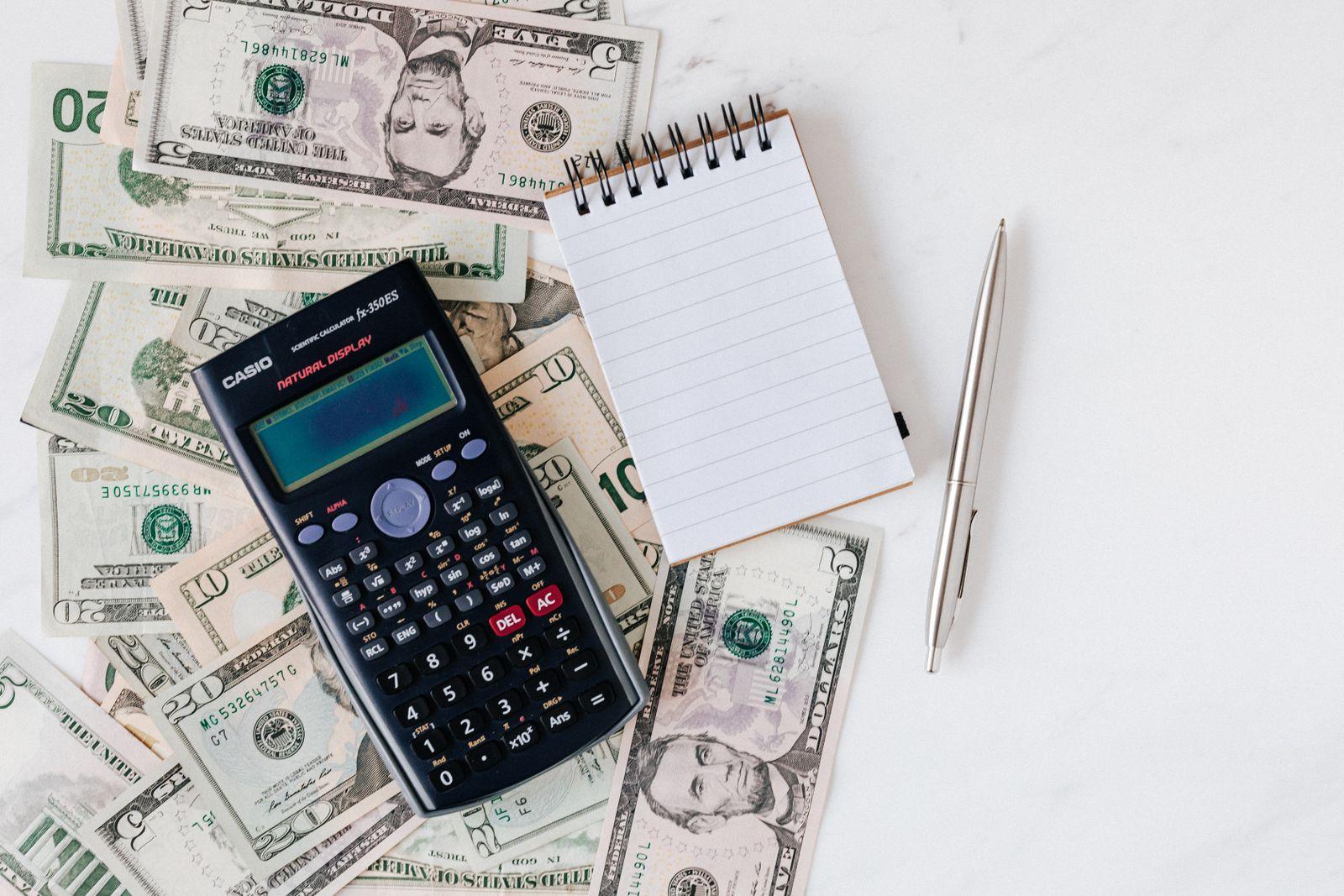 Una calculadora y un cuaderno sobre una mesa cubierta de billetes de dólar.